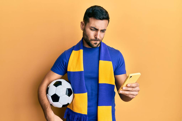 Young hispanic man football hooligan holding soccer ball using smartphone skeptic and nervous, frowning upset because of problem. negative person.  - 写真・画像