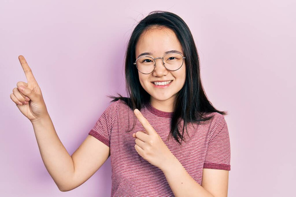 Fiatal kínai lány alkalmi ruhát és szemüveget visel mosolyog, és a kamerába néz, két kézzel és ujjal oldalra mutatva..  - Fotó, kép