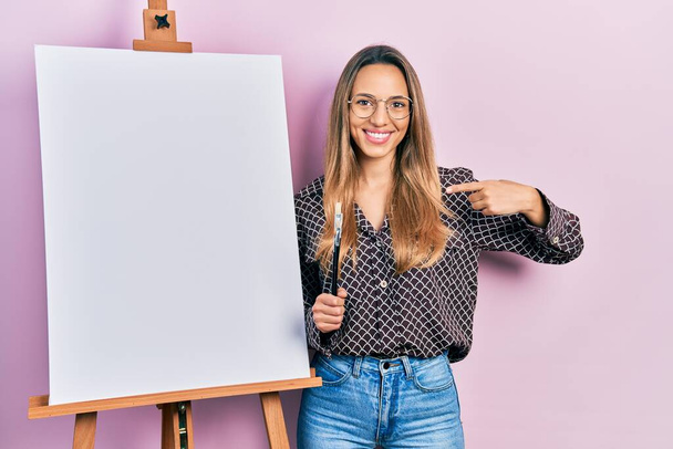 Belle femme hispanique debout près du peintre chevalet stand tenant pinceaux pointant du doigt à un sourire heureux et fier  - Photo, image