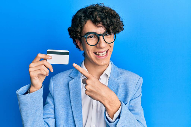 Ισπανόφωνος νεαρός κρατώντας πιστωτική κάρτα χαμογελώντας χαρούμενος δείχνοντας με το χέρι και το δάχτυλο  - Φωτογραφία, εικόνα