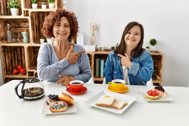 Семья матери и синдром Дауна дочь сидит дома едят завтрак весело с улыбкой лица указывая рукой и пальцем в сторону со счастливым и естественным выражением на лице  - Фото, изображение