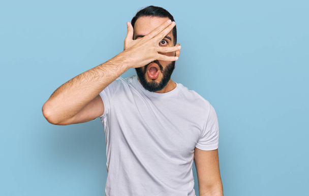 Young man with beard wearing casual white t shirt peeking in shock covering face and eyes with hand, looking through fingers with embarrassed expression.  - Fotoğraf, Görsel