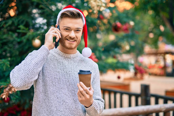 Young caucasian man wearing christmas hat talking on the smartphone and drinking coffee at the park - Photo, Image