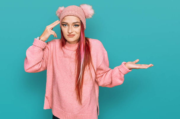 Young caucasian woman wearing casual clothes and wool cap confused and annoyed with open palm showing copy space and pointing finger to forehead. think about it.  - Foto, afbeelding