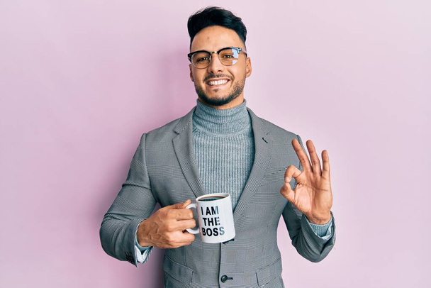 Young arab man drinking from i am the boss coffee cup doing ok sign with fingers, smiling friendly gesturing excellent symbol  - Zdjęcie, obraz