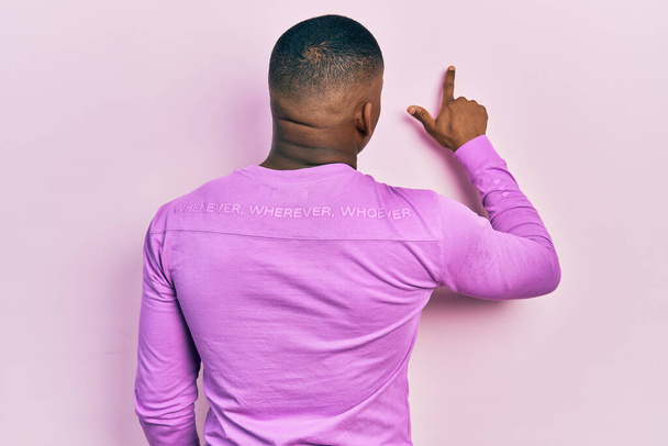 młody czarny człowiek noszenie casual różowy sweter pozowanie tył do przodu z palcem dłoni  - Zdjęcie, obraz