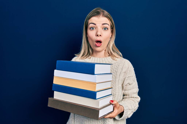 Beautiful caucasian woman holding a pile of books afraid and shocked with surprise and amazed expression, fear and excited face.  - Photo, image