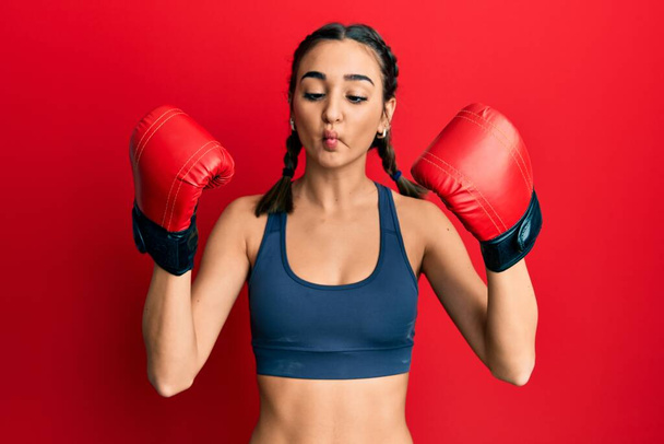 Young brunette girl using boxing gloves wearing braids making fish face with mouth and squinting eyes, crazy and comical.  - Foto, imagen