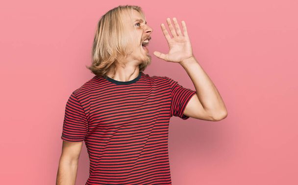 Homme caucasien aux cheveux longs blonds portant un t-shirt à rayures décontractées criant et criant haut et fort avec la main sur la bouche. concept de communication.  - Photo, image