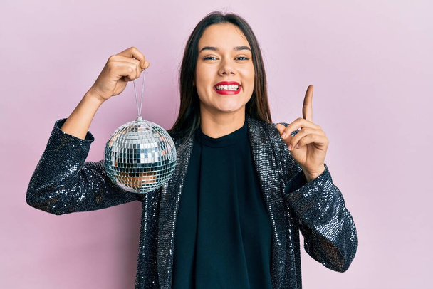 Jeune fille hispanique tenant une boule disco brillante souriant avec une idée ou une question pointant du doigt avec un visage heureux, numéro un  - Photo, image