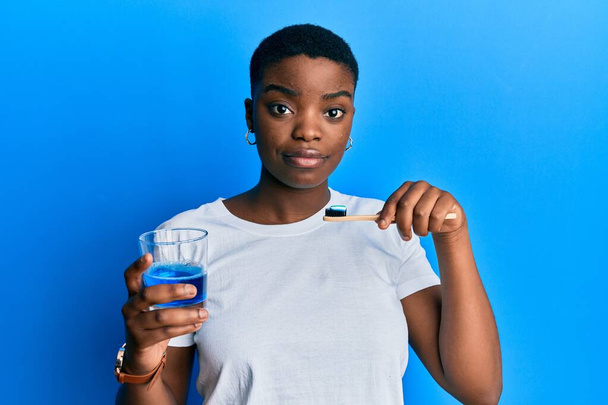 Genç Afro-Amerikalı kadın elinde bir bardak gargara ve diş fırçası tutuyordu. Temiz nefes almak için. Yüzünde ciddi bir ifadeyle rahatlamıştı. Basit ve doğal bir şekilde kameraya bakıyorsun..  - Fotoğraf, Görsel