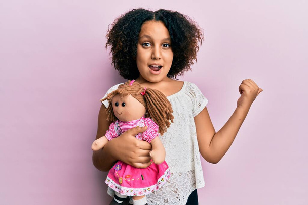 Jeune fille avec des cheveux afro tenant animal poupée jouet pointant pouce vers le côté souriant heureux avec bouche ouverte  - Photo, image