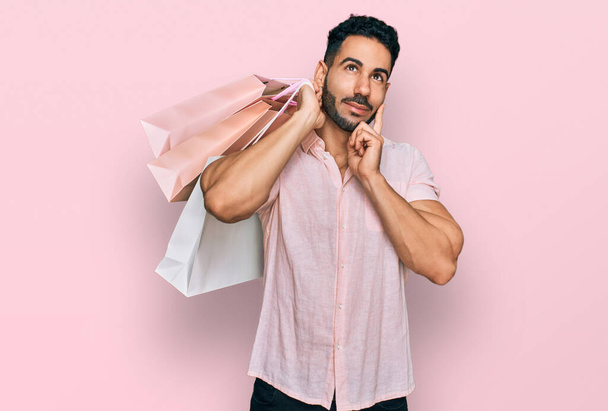 spanischer Mann mit Bart, der Einkaufstüten in der Hand hält, ernstes Gesicht, über Frage nachdenkend mit der Hand am Kinn, nachdenklich über verwirrende Idee  - Foto, Bild