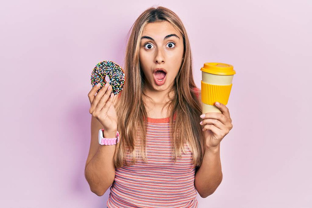 Beautiful hispanic woman eating doughnut and drinking coffee afraid and shocked with surprise and amazed expression, fear and excited face.  - Photo, Image