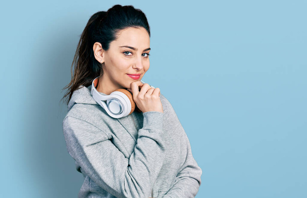 Young hispanic woman wearing gym clothes and using headphones smiling looking confident at the camera with crossed arms and hand on chin. thinking positive.  - Fotoğraf, Görsel