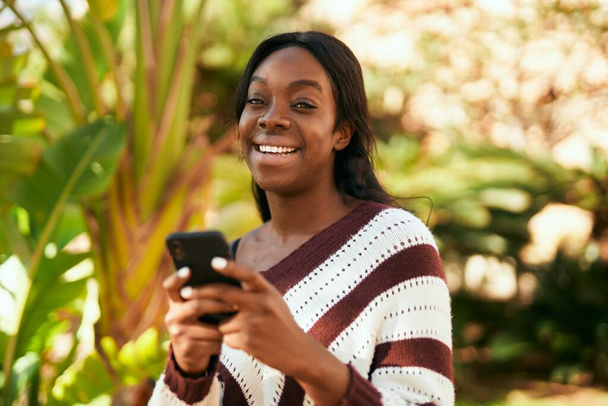 Młoda Afroamerykanka uśmiecha się szczęśliwa korzystając ze smartfona w parku. - Zdjęcie, obraz