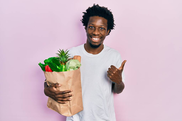 Jovem afro-americano segurando saco de papel com pão e mantimentos sorrindo feliz e positivo, polegar para cima fazendo excelente e sinal de aprovação  - Foto, Imagem