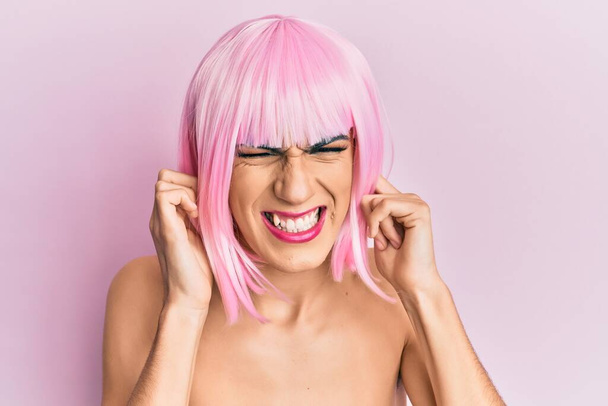 Młody mężczyzna, ubrany w kobietę, nosi różową perukę zakrywającą uszy palcami z irytującym wyrazem twarzy dla hałasu głośnej muzyki. koncepcja głucha.  - Zdjęcie, obraz