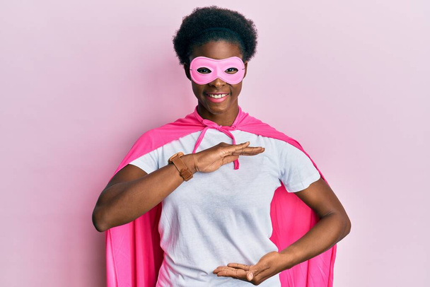 Young african american girl wearing superhero mask and cape costume gesturing with hands showing big and large size sign, measure symbol. smiling looking at the camera. measuring concept.  - Fotografie, Obrázek