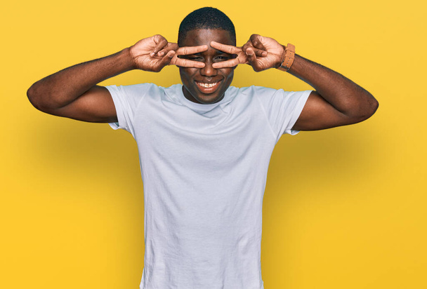 Jeune homme afro-américain portant un t-shirt blanc décontracté faisant symbole de paix avec les doigts sur le visage, souriant joyeux montrant la victoire  - Photo, image