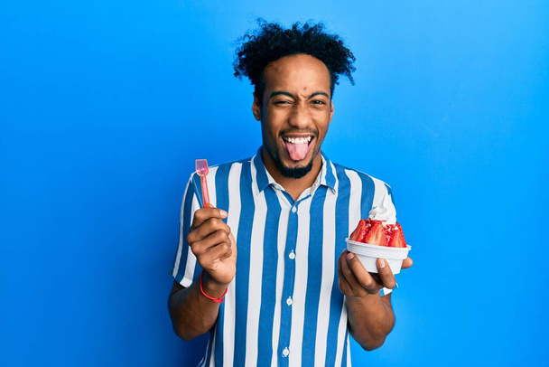 Jovem afro-americano com barba comendo sorvete de morango colando a língua feliz com expressão engraçada.  - Foto, Imagem