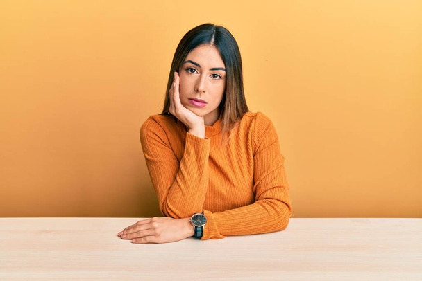 Junge hispanische Frau in lässiger Kleidung sitzt am Tisch und sieht müde und gelangweilt von Depressionen mit verschränkten Armen aus.  - Foto, Bild
