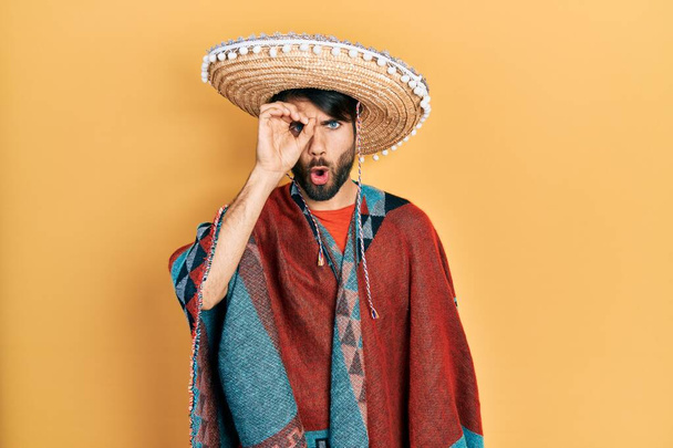 Νεαρός Ισπανός που κρατάει μεξικάνικο καπέλο κάνει καλή χειρονομία σοκαρισμένος με έκπληκτο πρόσωπο, με μάτια που κοιτούν μέσα από δάχτυλα. άπιστη έκφραση.  - Φωτογραφία, εικόνα