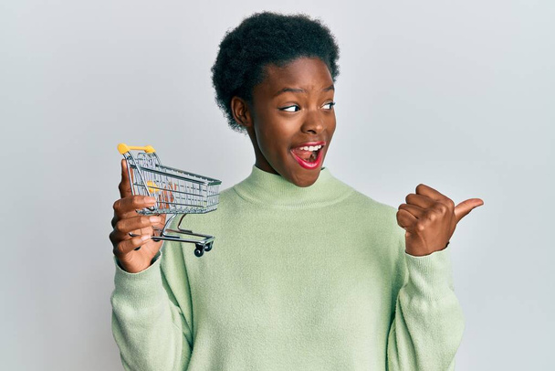 Young african american girl holding small supermarket shopping cart pointing thumb up to the side smiling happy with open mouth  - Foto, imagen