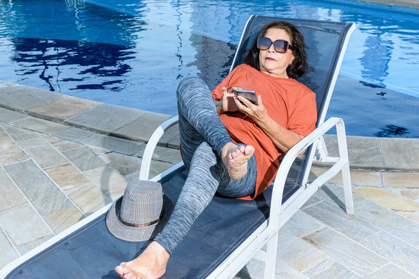 74 ετών γυναίκα κάθεται σε μια ξαπλώστρα και χρησιμοποιεί το smartphone της. - Φωτογραφία, εικόνα