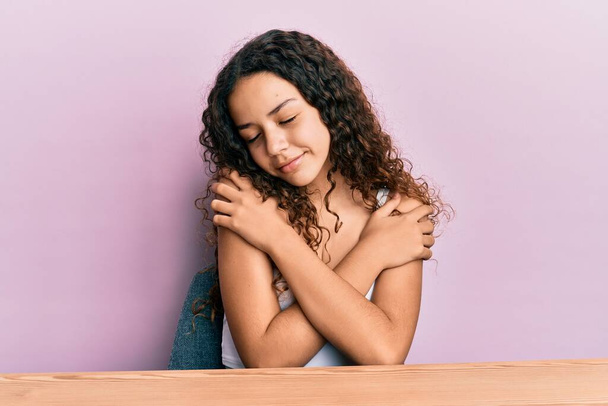 10代のヒスパニック系の女の子のカジュアル服をテーブルの上に座って自分自身幸せと肯定的な抱擁を着て、自信を笑顔。自己愛と自己ケア  - 写真・画像