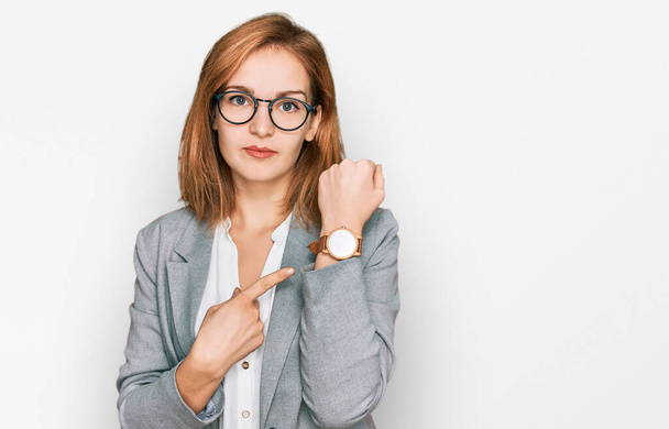 Молодая белая женщина в деловом стиле и в очках в спешке указывает на часы, нетерпение, смотрит в камеру с расслабленным выражением лица  - Фото, изображение