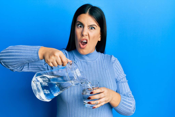 Όμορφη μελαχρινή γυναίκα ρίχνει νερό σε γυαλί στο πρόσωπο σοκ, αναζητούν δύσπιστοι και σαρκαστική, έκπληκτος με ανοιχτό στόμα  - Φωτογραφία, εικόνα