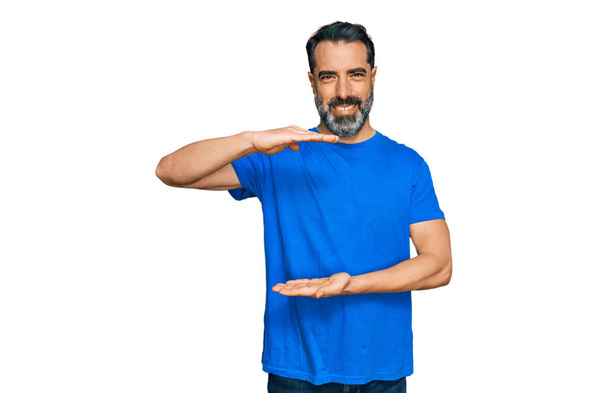 Man van middelbare leeftijd met baard in casual blauw t-shirt gebaren met wijzers met groot en groot maatbord, maatsymbool. glimlachend kijkend naar de camera. meetconcept.  - Foto, afbeelding