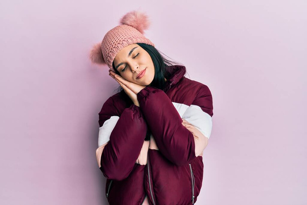 Młoda Latynoska kobieta w wełnianym zimowym swetrze i czapce śpi zmęczona snem i pozuje dłońmi razem, uśmiechając się zamkniętymi oczami.  - Zdjęcie, obraz