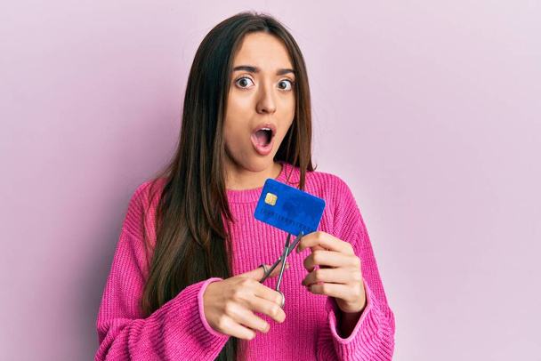 Young hispanic girl cutting credit card using scissors afraid and shocked with surprise and amazed expression, fear and excited face.  - Foto, immagini