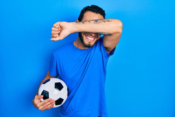 Uomo ispanico con la barba che tiene il pallone da calcio sorridente allegro che gioca a sbirciare una bua con le mani che mostrano la faccia. sorpreso ed uscito  - Foto, immagini