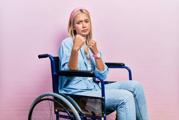 Beautiful blonde woman sitting on wheelchair ready to fight with fist defense gesture, angry and upset face, afraid of problem  - Foto, imagen