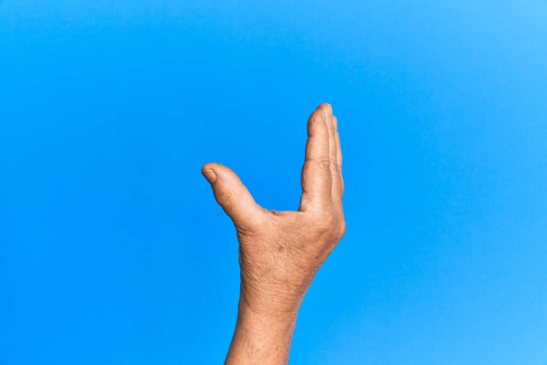 Mano de hombre hispano mayor sobre fondo azul aislado recogiendo y tomando lo invisible, sosteniendo el objeto con los dedos mostrando espacio  - Foto, imagen