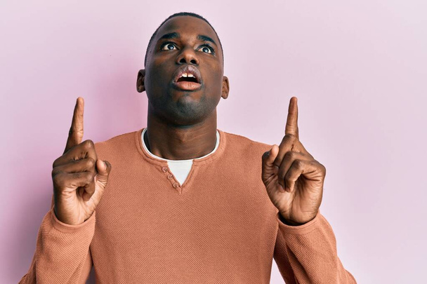 Junger afrikanisch-amerikanischer Mann in lässiger Kleidung staunt und überrascht und zeigt mit erhobenen Fingern und erhobenen Armen nach oben.  - Foto, Bild