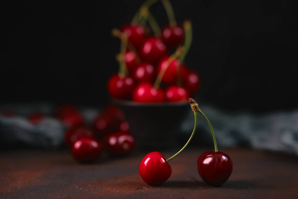 Όμορφο διπλό κεράσι σε πρώτο πλάνο και φρέσκα και γλυκά κόκκινα κεράσια σε μαύρο πήλινο μπολ σε σκούρο φόντο με σταγόνες νερού. εποχιακά φρούτα, λιχουδιά - Φωτογραφία, εικόνα