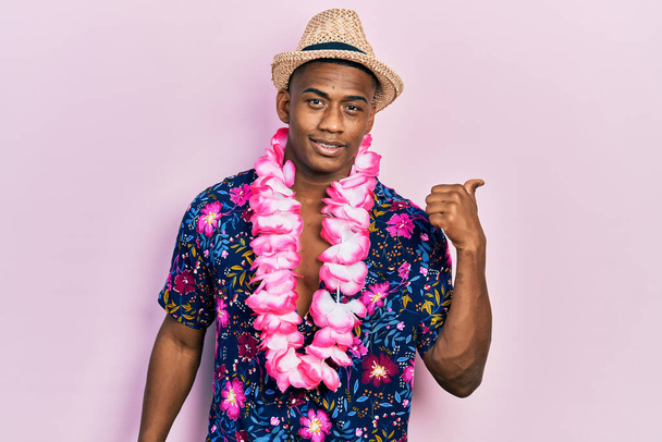 Fiatal fekete férfi nyári ingben és hawaii lei mosollyal boldog arccal néz, és feltartott hüvelykujjal oldalra mutat..  - Fotó, kép