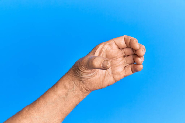 Ruka staršího hispánského muže přes modré izolované pozadí drží neviditelný objekt, prázdná ruka dělá výstřižek a uchopení gesto  - Fotografie, Obrázek