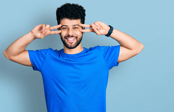 Молодой араб с бородой в повседневной синей футболке делает символ мира с пальцами на лице, улыбаясь весело показывая победу  - Фото, изображение