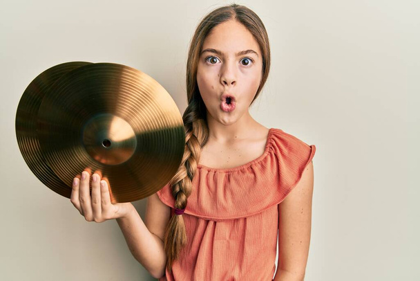 Belle petite fille brune tenant des plaques de cymbales dorées effrayées et étonnées avec la bouche ouverte pour la surprise, visage incrédule  - Photo, image