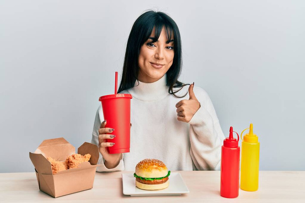 Young brunette woman with bangs eating a tasty classic burger with ketchup and mustard smiling happy and positive, thumb up doing excellent and approval sign  - Foto, imagen