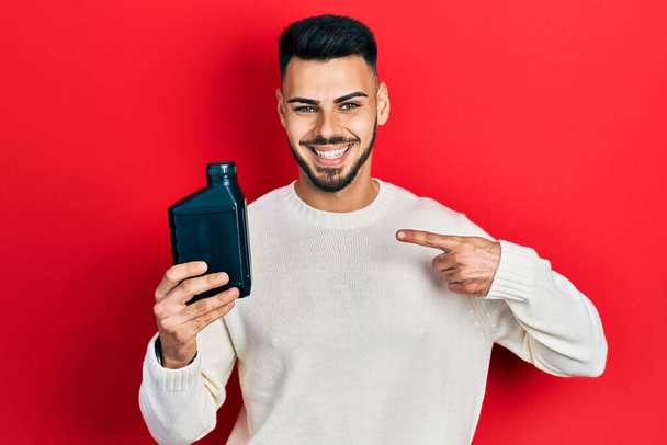 Young hispanic man with beard holding motor oil bottle smiling happy pointing with hand and finger  - Fotoğraf, Görsel