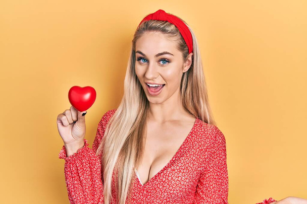Young caucasian woman holding red heart celebrating achievement with happy smile and winner expression with raised hand  - Photo, Image