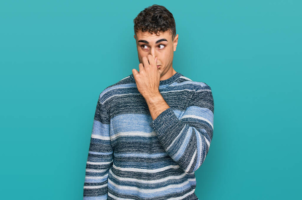 Ισπανόφωνος νεαρός που φοράει καθημερινά ρούχα μυρίζοντας κάτι βρωμερό και αηδιαστικό, ανυπόφορη μυρωδιά, κρατώντας ανάσα με δάχτυλα στη μύτη. άσχημη μυρωδιά  - Φωτογραφία, εικόνα