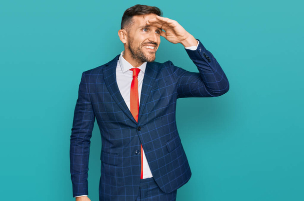 Handsome man with beard wearing business suit and tie very happy and smiling looking far away with hand over head. searching concept.  - Foto, Imagen