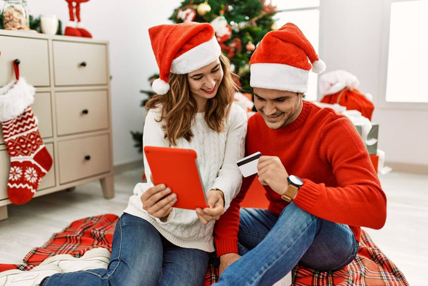 Молодая испаноязычная пара в рождественской шляпе с помощью тачпада и кредитной карты сидит на полу дома. - Фото, изображение